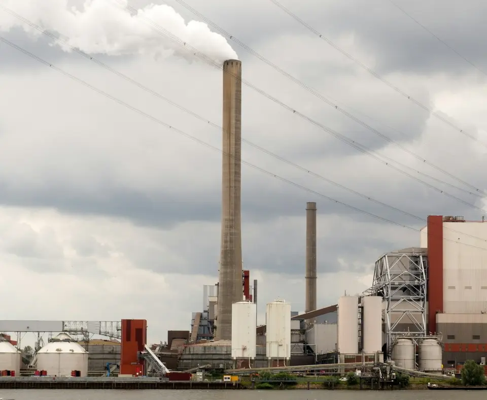 De Amercentrale bij Geertruidenberg: een hoge schoorsteen met een witte rookpluim, silo's en een ketelhuis aan het water, onder hoogspanningskabels en een grijze lucht